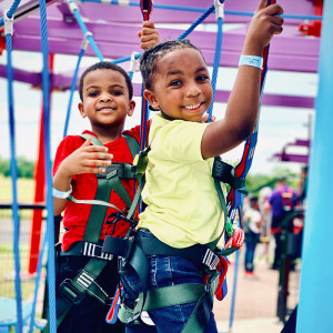 Two children are smiling and climbing a ropes course at the family fun center, both secured in safety harnesses. One dons a yellow shirt while the other sports red, set against colorful structures and a clear sky. It's an adventure rivaling a perfect day at Spare Time Texas or even bowling with friends.