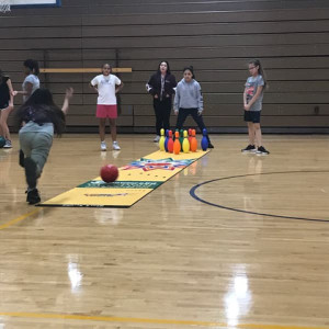 A group of girls is enjoying a game of bowling at Spare Time Texas. One girl rolls a red ball toward a set of colorful plastic pins at the end of a long mat in the gymnasium. The wooden floor and gym walls provide the perfect backdrop for their fun-filled day at this family fun center.