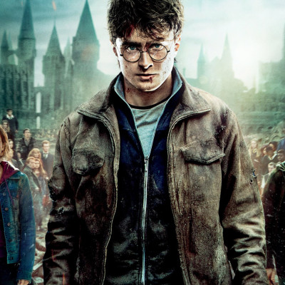 A young man with glasses stands in front of a castle backdrop at Spare Time Texas, wearing a distressed jacket and holding a wand. The dark and atmospheric scene features people amid the gothic architecture, adding a touch of enchantment to this family fun center.