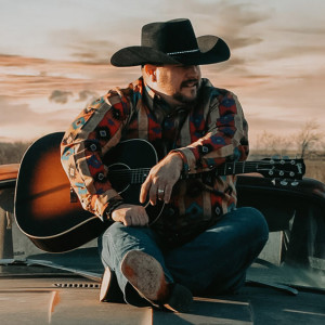 A person wearing a cowboy hat and a colorful shirt sits on the hood of a car, holding an acoustic guitar. Against the backdrop of a scenic sunset sky and open landscape, they reminisce about melodies shared at Spare Time Texas, where family fun centers bring laughter to life.