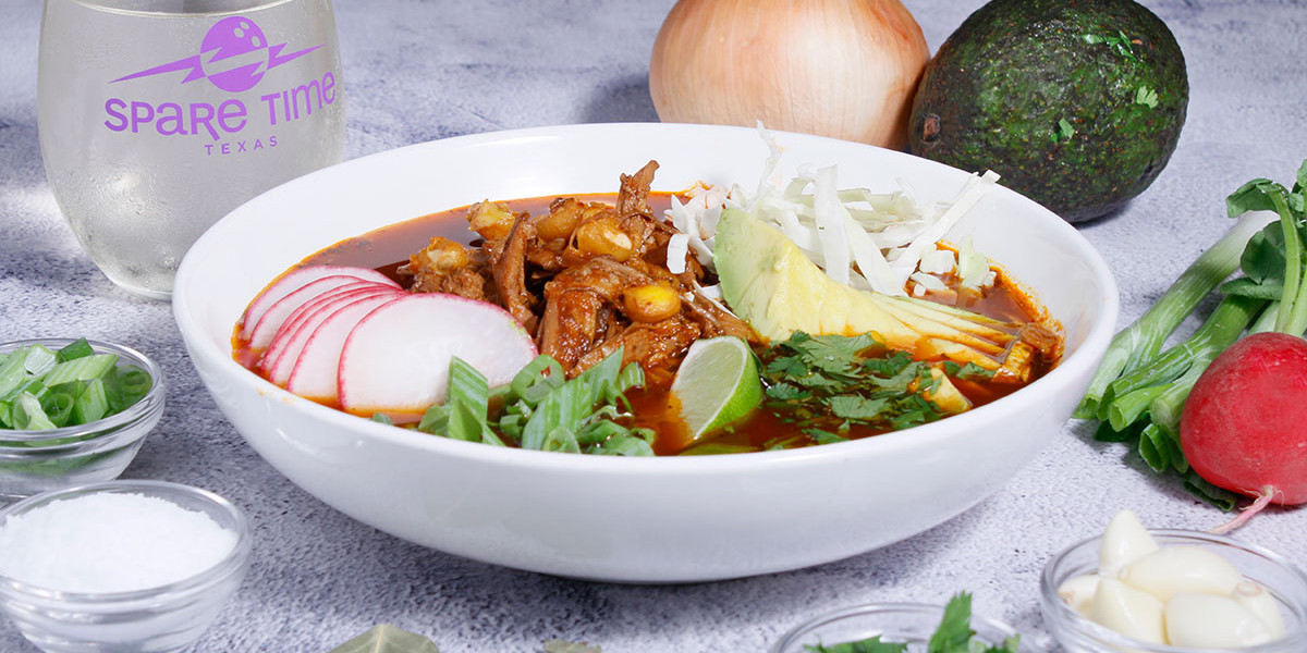 A bowl of Mexican pozole garnished with sliced radishes, avocado, lime, cabbage, and cilantro sits on the table. Nearby are an avocado and onions next to a glass branded with "Spare Time Texas," perfect for a post-bowling meal at the family fun center.