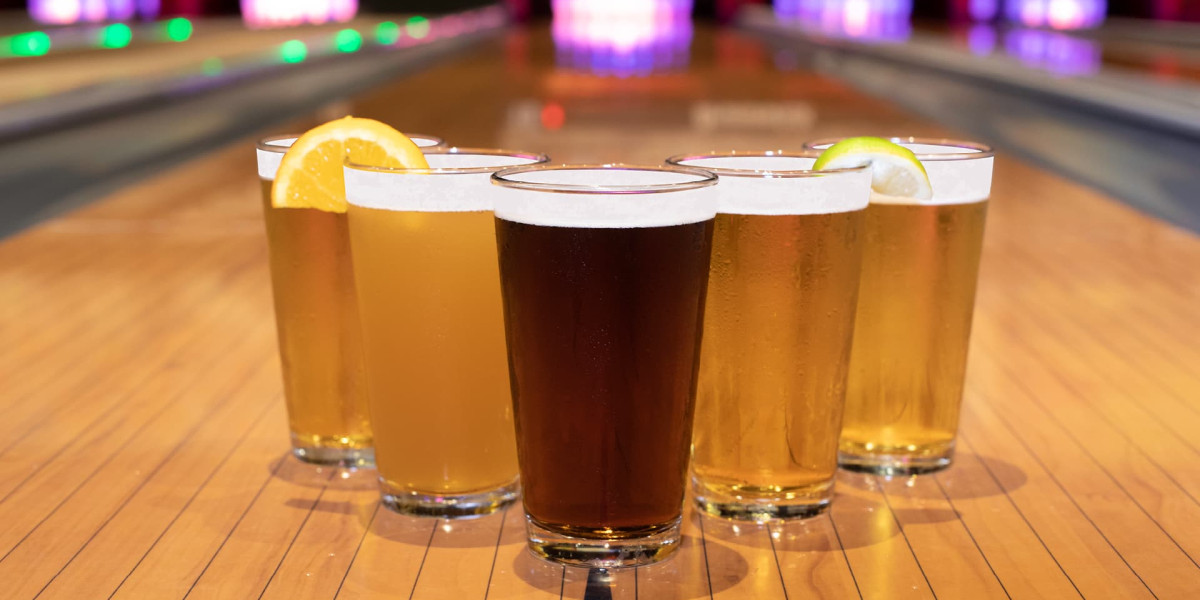 Five pint glasses filled with different types of beer, some garnished with orange and lime slices, are placed on a Spare Time Texas bowling lane. The family fun center's bowling alley is brightly lit in the background, creating a lively atmosphere perfect for friends and family.