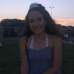 A person with long hair smiles while sitting outdoors on the grass during sunset, wearing a blue and white checkered dress. Nearby, scattered chairs hint at earlier family fun outside of Spare Time Texas, where laughter from bowling lanes still echoes in the background.