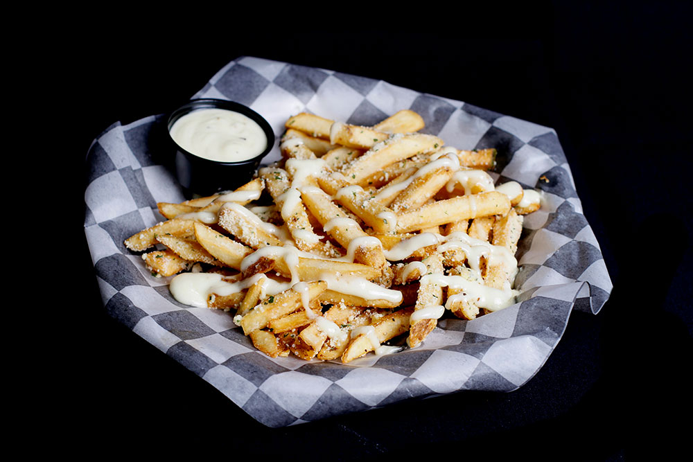 At Spare Time Texas, enjoy a basket of crinkle-cut French fries topped with melted cheese and garnished with seasonings. Perfect for sharing during a fun game of bowling, this treat comes with a small cup of creamy dipping sauce on black-and-white checkered paper.