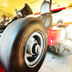 Close-up of a red go-kart with a large front tire in focus at Spare Time Texas, where bowling and racing collide for ultimate family fun. The background is blurred, suggesting speed or motion. The steering wheel and seat are visible, with sunlight streaming through a yellowish backdrop.