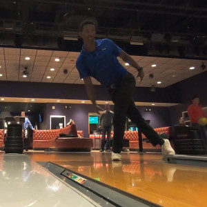 At Spare Time Texas, a person releases the bowling ball in mid-action down the polished lane. The dimly lit alley buzzes with excitement, while groups enjoy time together on red couches in the background of this vibrant family fun center.
