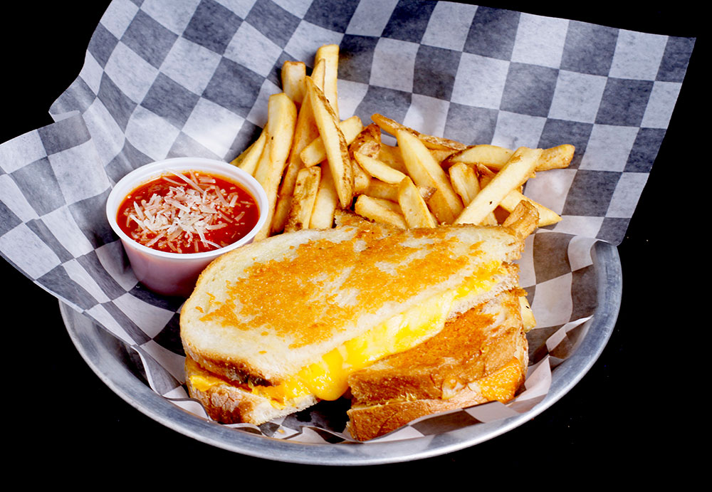 A grilled cheese sandwich with melted cheese is served on a checkered paper-lined plate, accompanied by golden French fries at Spare Time Texas. A small cup of marinara sauce topped with grated cheese sits beside them, making it the perfect treat after a fun-filled day at the family fun center.