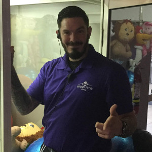 Man smiling and giving a thumbs-up, wearing a purple shirt and earpiece, stands near a claw machine filled with plush toys at Spare Time Texas. Perfect for family fun, this spot offers everything from bowling to arcade games for an unforgettable day of entertainment.