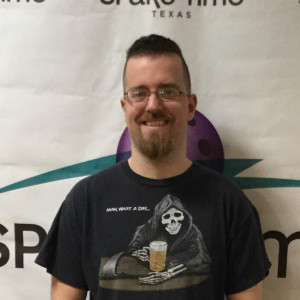 A smiling person with glasses and a beard stands in front of a banner at Spare Time Texas. They're wearing a t-shirt depicting a grim reaper holding a beer, along with the words "Have What a Die," capturing the playful spirit of this family fun center's bowling alley.