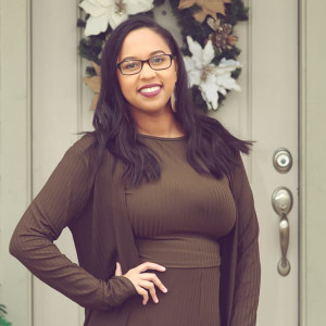 A woman with long dark hair and glasses stands smiling in front of a door decorated with a floral wreath. Dressed in a dark green dress and cardigan, she poses with one hand on her hip, ready for an evening of bowling at the Spare Time Texas family fun center.