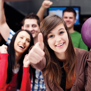 At Spare Time Texas, a group of four young adults celebrates with smiles and thumbs up at the family fun center's bowling alley. The woman in the foreground holds a purple ball, while others cheer joyfully in the background, making every strike an exciting moment.