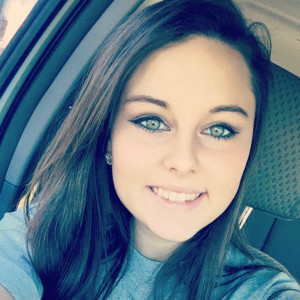 A young woman with long brown hair, wearing a gray shirt, is smiling while sitting inside a car, perhaps heading to Spare Time Texas for some family fun. Her bright eyes and eyeliner accentuate her excitement. The car seat is visible in the background.