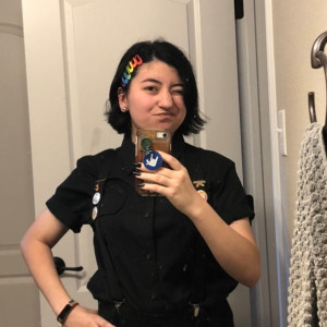 A person with short black hair takes a selfie in front of a mirror at Spare Time Texas. They’re wearing a black shirt with colorful clips in their hair and a playful expression, ready for some bowling. Their phone case features a blue sticker, embodying the spirit of the family fun center.