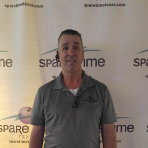 A person wearing a gray polo shirt with a logo stands in front of a vibrant backdrop featuring "Spare Time Texas" and a bowling graphic, highlighting the excitement of this family fun center. They have short hair and are equipped with an earpiece.