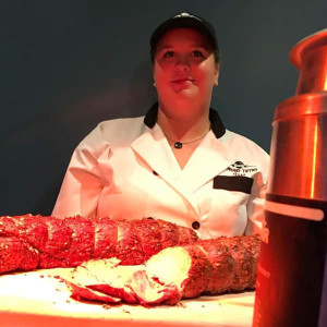 A chef in uniform sits behind a wooden cutting board with a large piece of seasoned meat, partially sliced. The image is warmly lit, highlighting the textures of the meat. Perfect for those planning a day at Spare Time Texas, this scene adds gourmet flair to your family fun center outing.