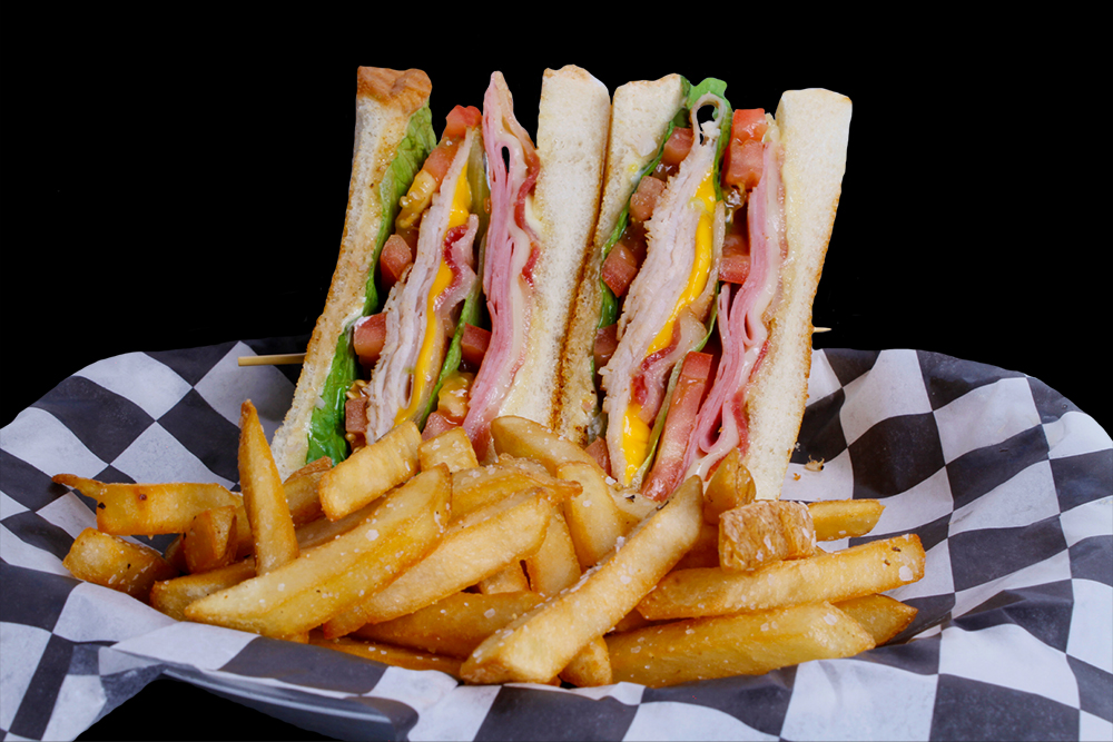 A club sandwich with layers of ham, lettuce, tomato, and cheese is cut into quarters and served on a black and white checkered paper at Spare Time Texas. Golden crispy fries are placed in front of the sandwich, making it perfect for enjoying between rounds at the family fun center.