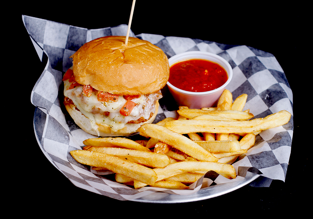 A cheeseburger with tomato and a wooden pick, served on checkered paper alongside crispy fries and a small cup of red sauce, awaits you on a metal plate at Spare Time Texas. It's the perfect fuel for an action-packed day of bowling at this vibrant family fun center.