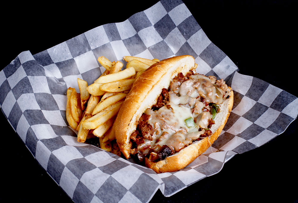 At Spare Time Texas, savor a delicious cheesesteak sandwich with melted cheese, steak, and peppers in a hoagie roll, served on checkered paper alongside golden French fries. It's the perfect way to refuel after some family fun at the bowling lanes.