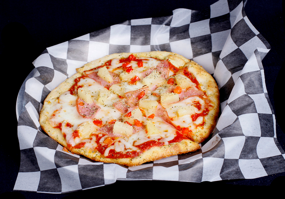 A Hawaiian pizza with ham and pineapple rests on black and white checkered paper at Spare Time Texas. The thin, golden-brown crust pairs perfectly with melted cheese and tomato sauce, making it an ideal treat to enjoy in their lively family fun center.