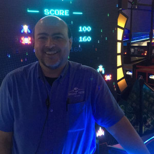 A person smiling and standing near an arcade game machine at Spare Time Texas. The game screen in the background shows a score of 160. The person, in a blue shirt, is illuminated by dim blue and neon lights, capturing the vibrant atmosphere of this family fun center.