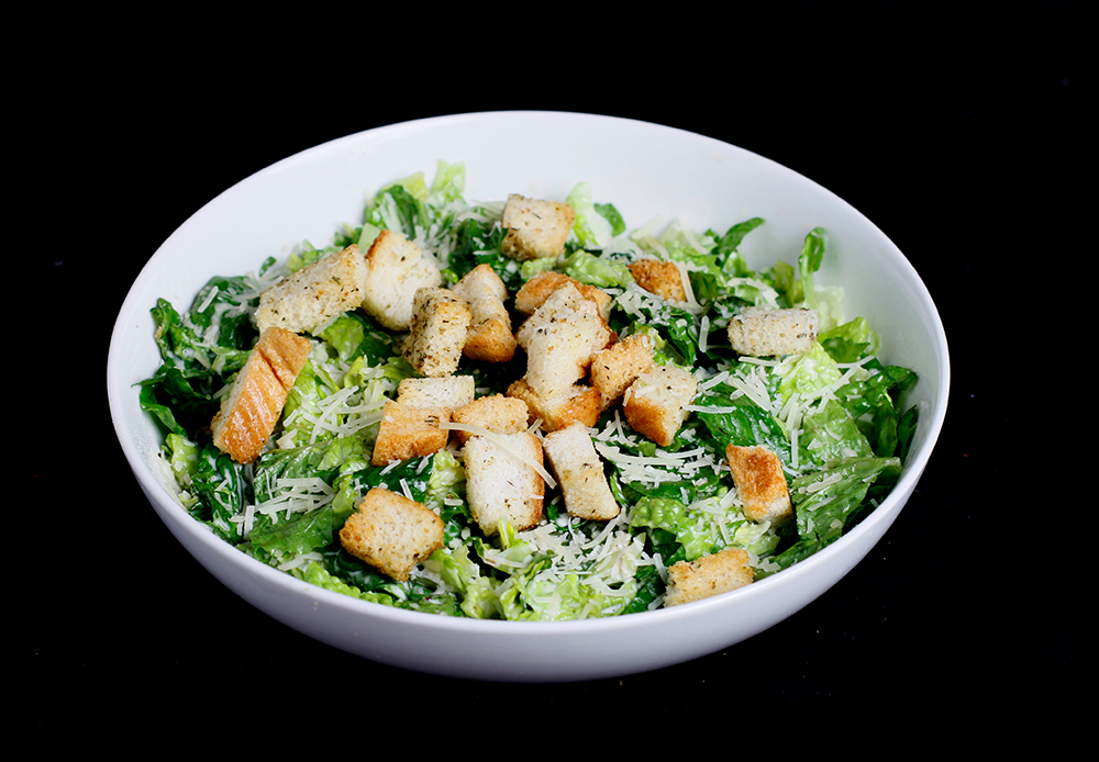 A bowl of Caesar salad with romaine lettuce, croutons, and grated Parmesan cheese takes center stage against a black background, reminiscent of a perfect evening at Spare Time Texas, where delicious dining meets the excitement of a family fun center.