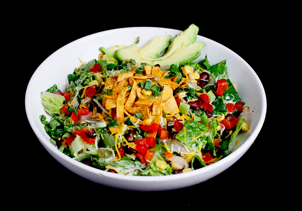 A white bowl filled with a mixed salad featuring lettuce, chopped tomatoes, black beans, corn, avocado slices, and grated cheese is topped with crispy tortilla strips and green onions. This vibrant dish is perfect for sharing at a family fun center like Spare Time Texas.