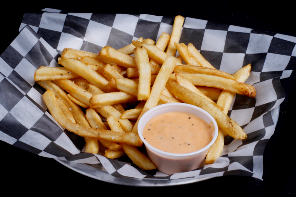 A basket of golden-brown french fries on black-and-white checkered paper, accompanied by a small cup of dipping sauce, makes for a perfect snack at Spare Time Texas. Enjoy your fries between bowling games or while exploring the family fun center's attractions.