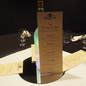 A menu titled "A Taste of Spain" is displayed upright with the help of a wine bottle on a dining table at Spare Time Texas, the ultimate family fun center. The menu includes Gazpacho Soup, Traditional Tapas, Seafood Paella, and Crema Catalana. Rolled napkins and glasses are placed nearby—perfect after some bowling fun!.
