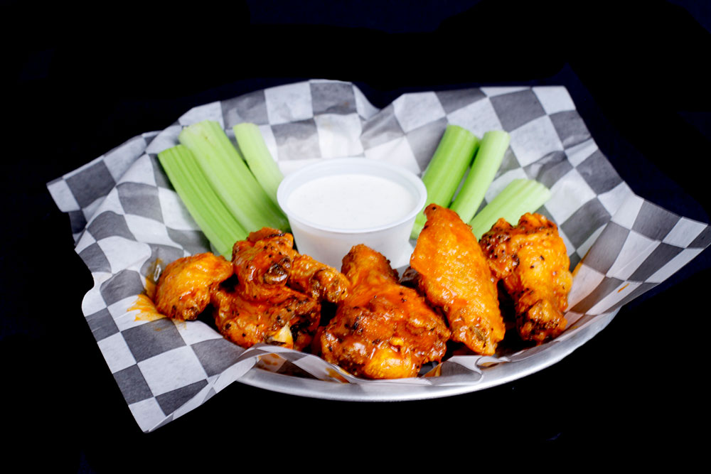 A plate with buffalo chicken wings, celery sticks, and a small cup of ranch dressing sits on a checkered paper lining at the Spare Time Texas family fun center, perfect for fueling up before hitting the bowling lanes.