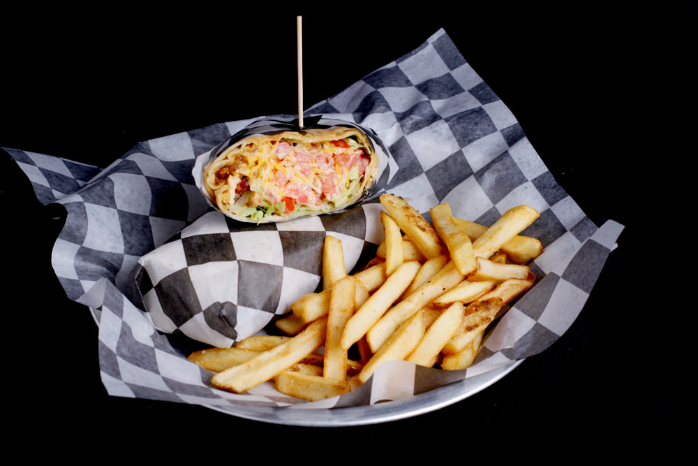 A basket lined with black and white checkered paper contains a sliced wrap filled with colorful ingredients and a serving of golden French fries, perfectly set against a black background, reminiscent of the vibrant vibes at Spare Time Texas or any lively family fun center.