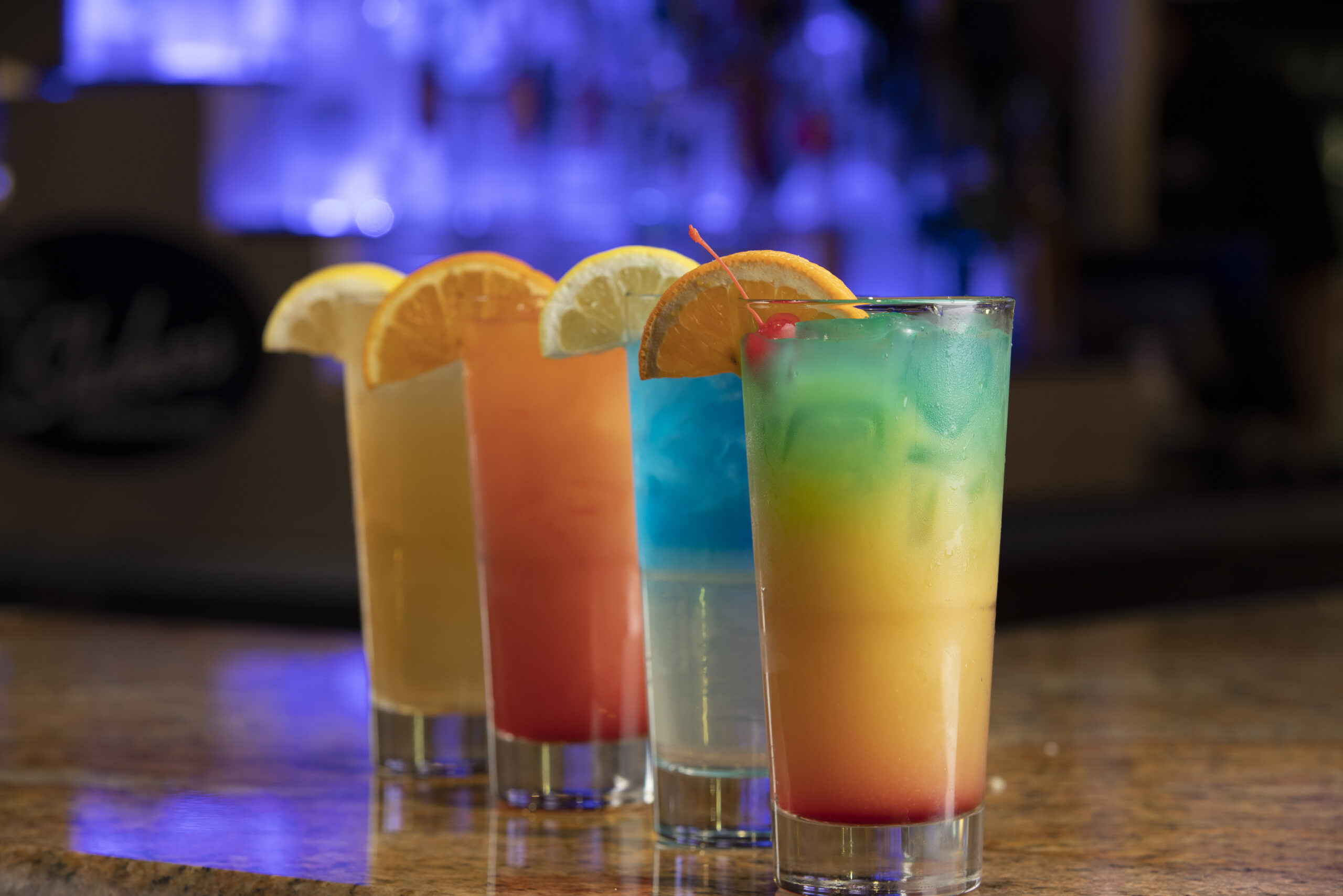 Four colorful cocktails in glasses on a wooden table, each garnished with a lemon slice. In the backdrop of Spare Time Texas, a dimly lit bar with a blue glow complements these vibrant drinks, from red to blue to orange. Perfect for unwinding after exciting rounds of bowling at this family fun center.