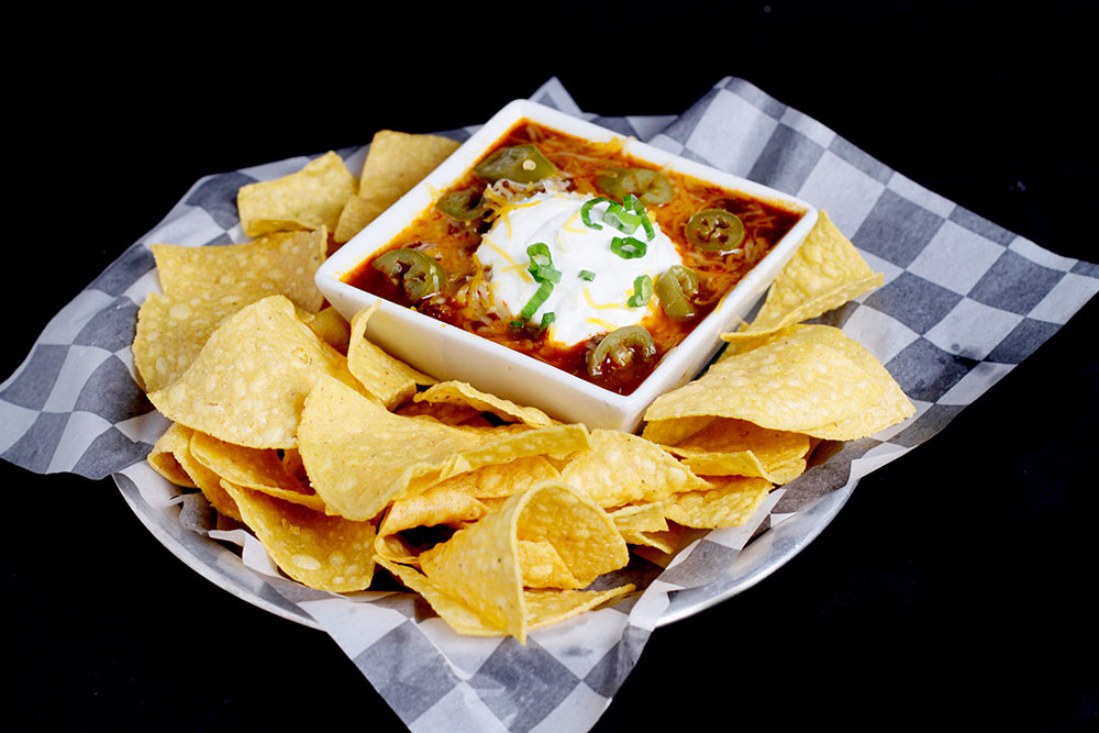 At Spare Time Texas, a plate of tortilla chips surrounds a square bowl of chili topped with sour cream, sliced jalapeños, and green onions, set on checkered paper. It's the perfect snack to enjoy at this family fun center after an exciting game of bowling.