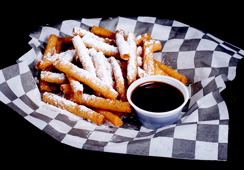 At Spare Time Texas, a basket of churros dusted with powdered sugar sits on black and white checkered paper. Perfect for sharing at this vibrant family fun center, it comes with a small cup of chocolate dipping sauce, making it an irresistible treat after a game of bowling.
