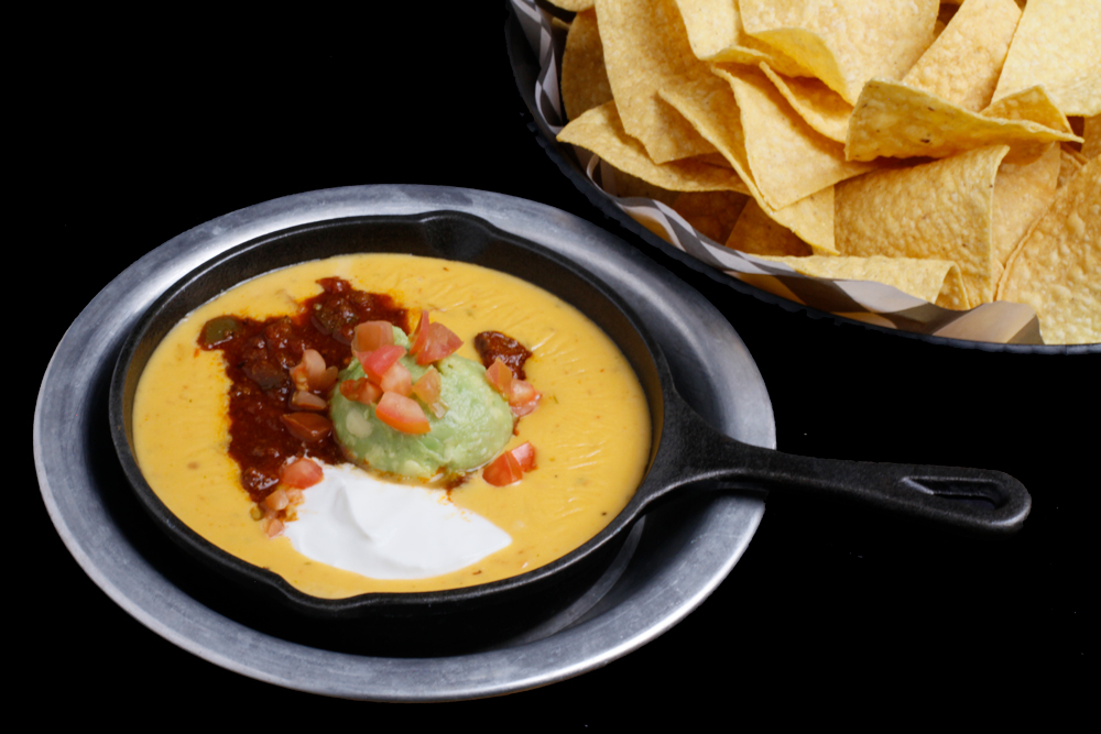 At Spare Time Texas, enjoy a skillet with melted cheese dip topped with green guacamole, red salsa, and a dollop of white sour cream. Perfect for sharing while taking a break from bowling or exploring the family fun center, with a bowl of yellow corn tortilla chips on the side.