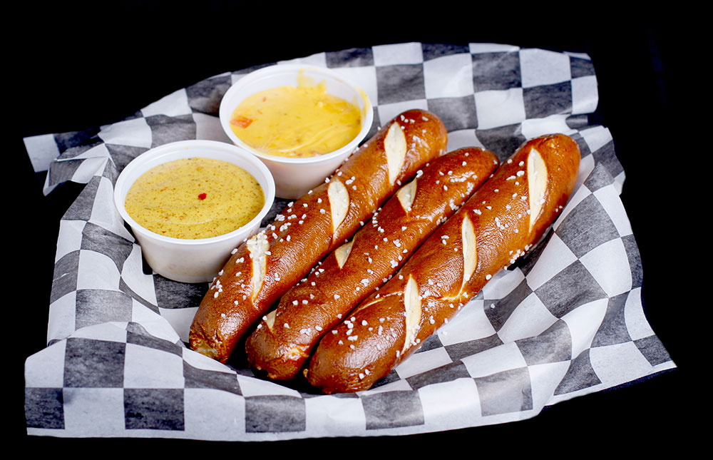 Three soft pretzel sticks with salt rest on a black and white checkered sheet at Spare Time Texas, served alongside two small containers of yellow mustard and cheese sauce. Enjoy this delicious snack while experiencing the excitement of our family fun center or during a friendly game of bowling.