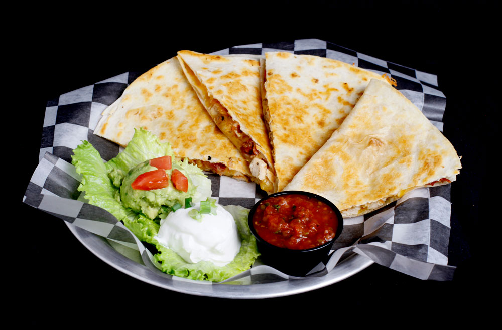 A plate with sliced quesadillas on black-and-white checkered paper at the Spare Time Texas family fun center. It’s accompanied by small portions of guacamole, sour cream, and salsa, garnished with lettuce and a few diced tomatoes—perfect for enjoying after a friendly game of bowling.
