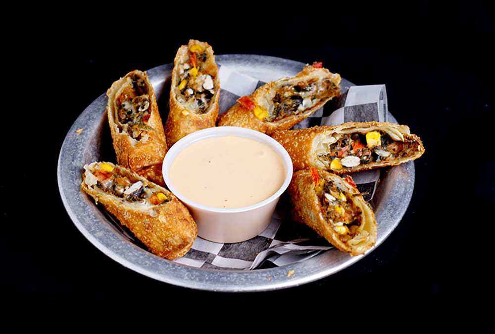A metal plate with crispy stuffed eggrolls arranged around a small cup of creamy dipping sauce on a black background, resembling the delightful snacks enjoyed at Spare Time Texas after an exciting day in the family fun center. The sliced eggrolls reveal a colorful filling of vegetables and beans.