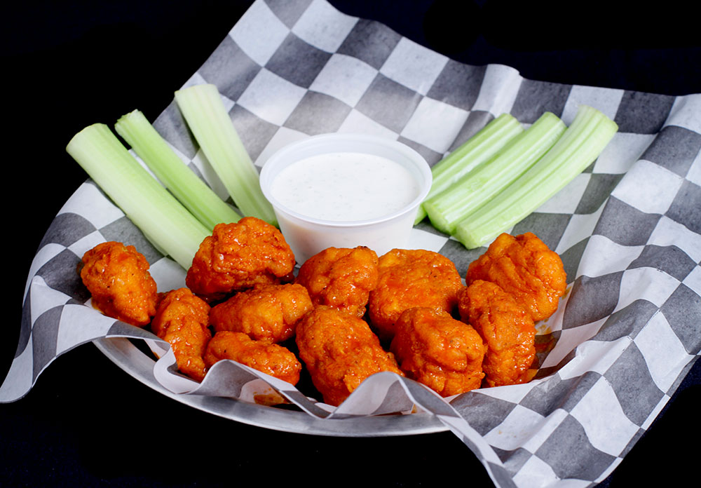 A plate of spicy buffalo wings drenched in red-orange sauce, accompanied by celery sticks and a cup of ranch dressing, awaits on checkered paper—perfect for fueling up at Spare Time Texas, where family fun and bowling excitement collide.