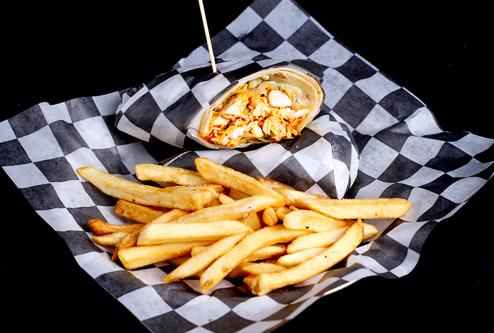 At Spare Time Texas, enjoy a delicious wrap filled with chicken and other ingredients, partially sliced and secured with a toothpick on black and white checkered paper. Pair it with golden-brown French fries for the perfect snack during your family fun center bowling adventure.