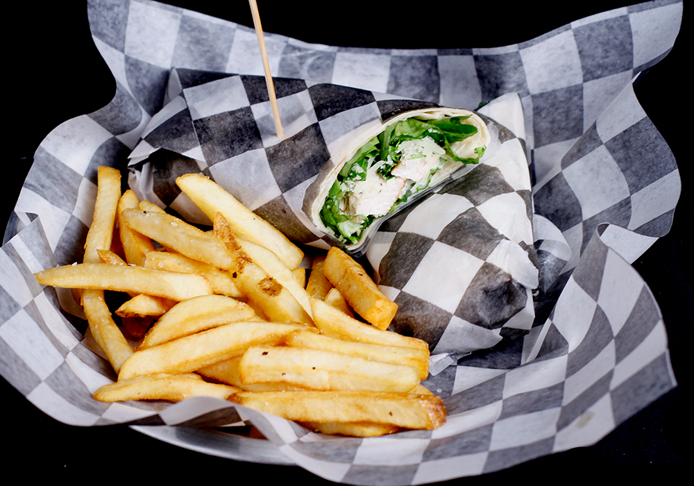 A basket lined with checkered paper holds a wrap filled with chicken and lettuce, sliced in half and secured with a toothpick. Perfect for enjoying between turns at the bowling lanes during your visit to Spare Time Texas, it's accompanied by a serving of golden, crispy fries.