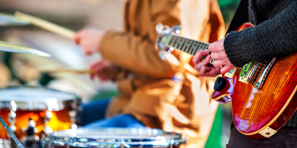 In a lively scene reminiscent of a family fun center, two musicians perform outdoors. The focus is on a guitarist in the foreground with a brightly colored electric guitar, while in the background, a drummer plays his set. Both are dressed casually, much like you’d dress for an outing at Spare Time Texas.