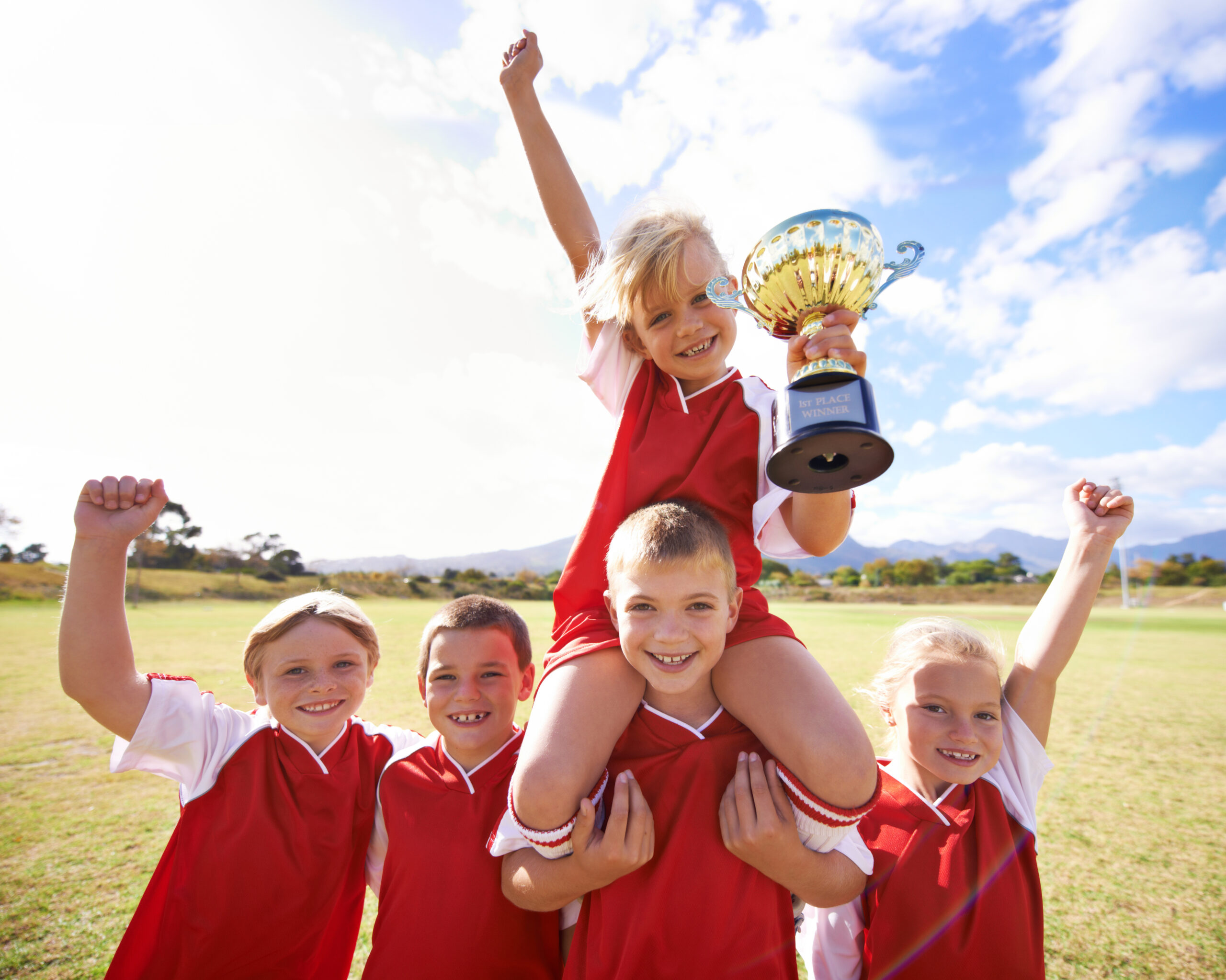 A group of smiling children in red sports uniforms celebrate on a grassy field, reminiscent of a day at Spare Time Texas. One child sits on another's shoulders, triumphantly holding a trophy, while the others raise their arms in victory. The sky is clear with a few clouds.