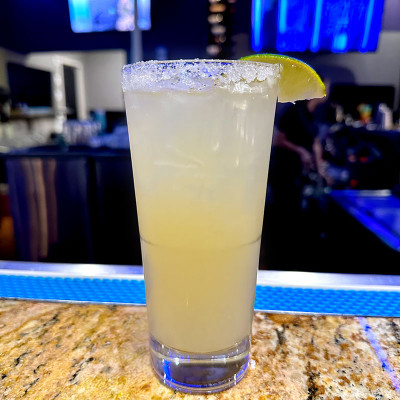 A tall glass of margarita with a salted rim and a lime wedge garnish sits on the granite bar countertop at Spare Time Texas. The dimly lit bar area, bathed in blue and purple lights, offers a stylish retreat after an exciting evening of bowling at this family fun center.