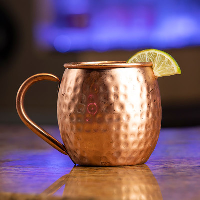 A copper mug with a hammered texture sits on a countertop at Spare Time Texas. A slice of lime is perched on the rim, while blurred hues of blue and purple in the background evoke the vibrant energy of this family fun center.