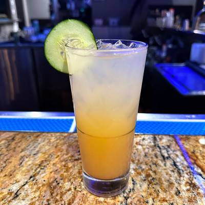 A tall glass filled with a refreshing iced drink topped with a lime slice rests on the granite countertop at Spare Time Texas. The drink's golden to pale yellow gradient catches the dim light, hinting at the lively atmosphere of this popular family fun center, where bowling lanes await in the background.