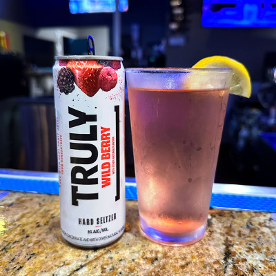 A can of Truly Wild Berry hard seltzer sits next to a glass filled with the drink, garnished with a lemon wedge, all set on a granite countertop. The backdrop evokes the lively ambiance of Spare Time Texas bowling lanes, perfect for capturing moments of family fun and refreshment.