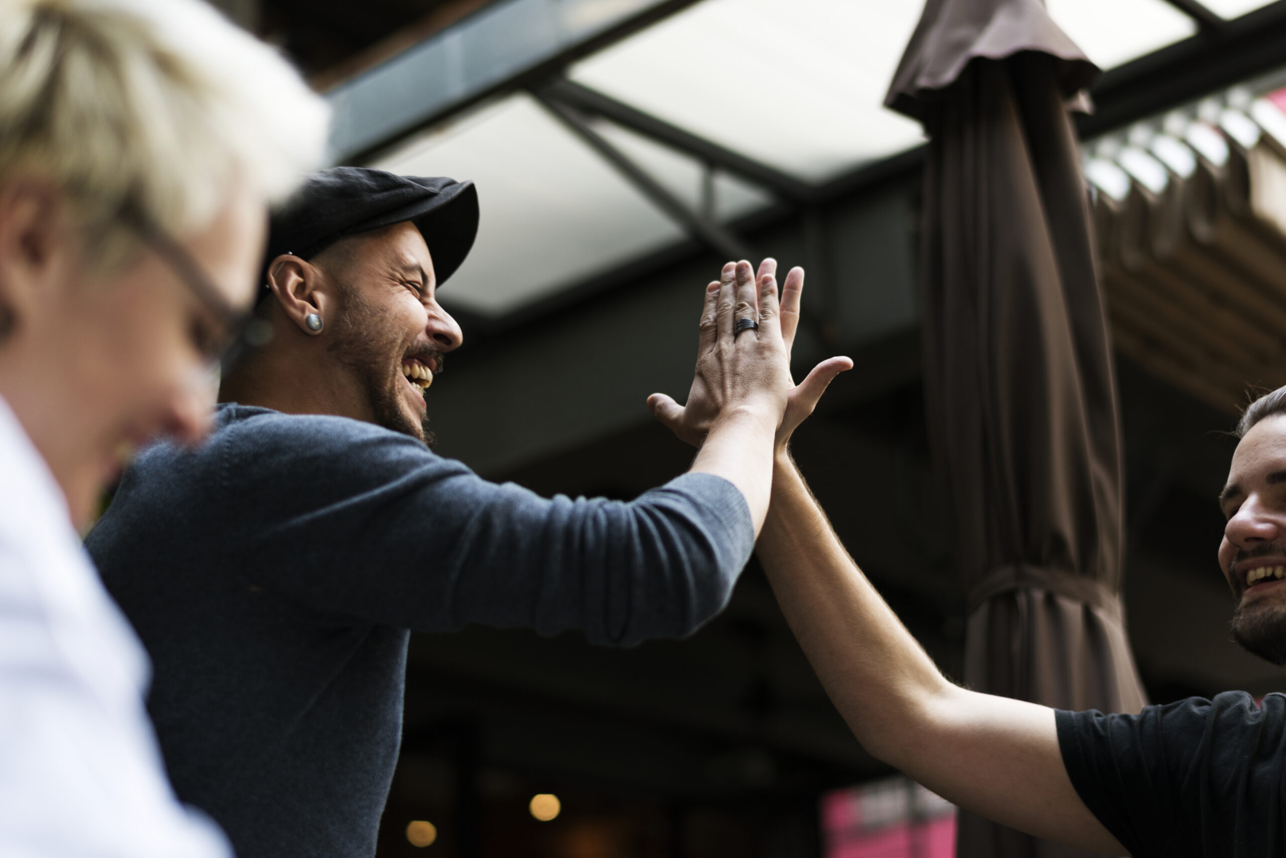 Two people smiling and giving each other a high five outdoors, with one wearing a cap while the other is off-camera at Spare Time Texas. Another person is slightly blurred in the foreground. They appear to be enjoying a light-hearted moment at this exciting family fun center.
