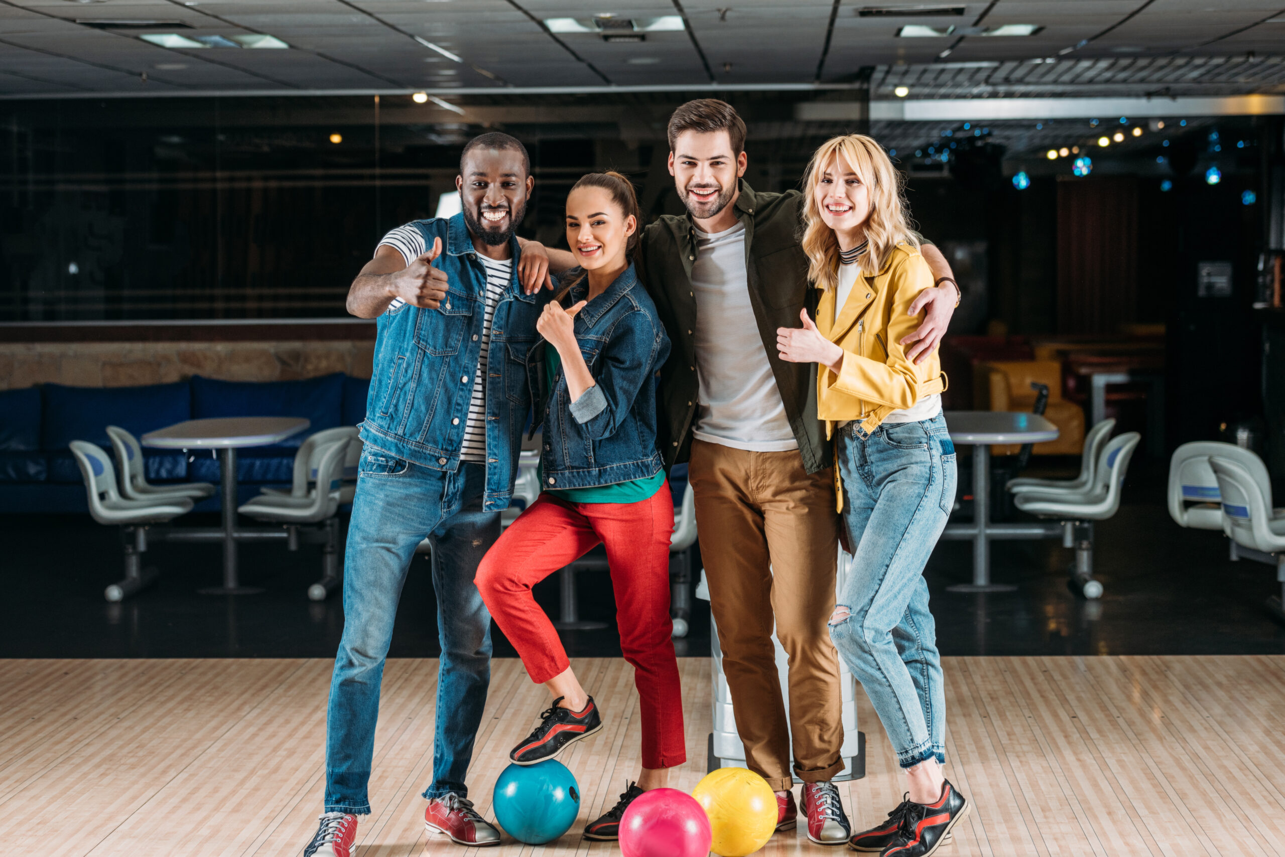 Four friends in casual clothes stand on a bowling lane at Spare Time Texas, smiling with arms around each other. Three bowling balls rest on the floor before them. The backdrop reveals more lanes and seating, capturing the essence of this lively family fun center.