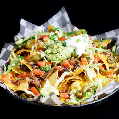 At Spare Time Texas, dive into a basket of nachos topped with melted cheese, ground meat, diced tomatoes, jalapenos, shredded lettuce, guacamole, sour cream, and chopped green onions—all perfect for sharing during a thrilling bowling game or family fun center adventure on our checkered paper liner.