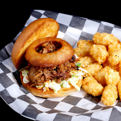 Enjoy a pulled pork sandwich topped with a crispy onion ring and coleslaw on a toasted bun, served with golden-brown tater tots at Spare Time Texas. It's the perfect meal to fuel your fun at this family-friendly bowling and entertainment center.